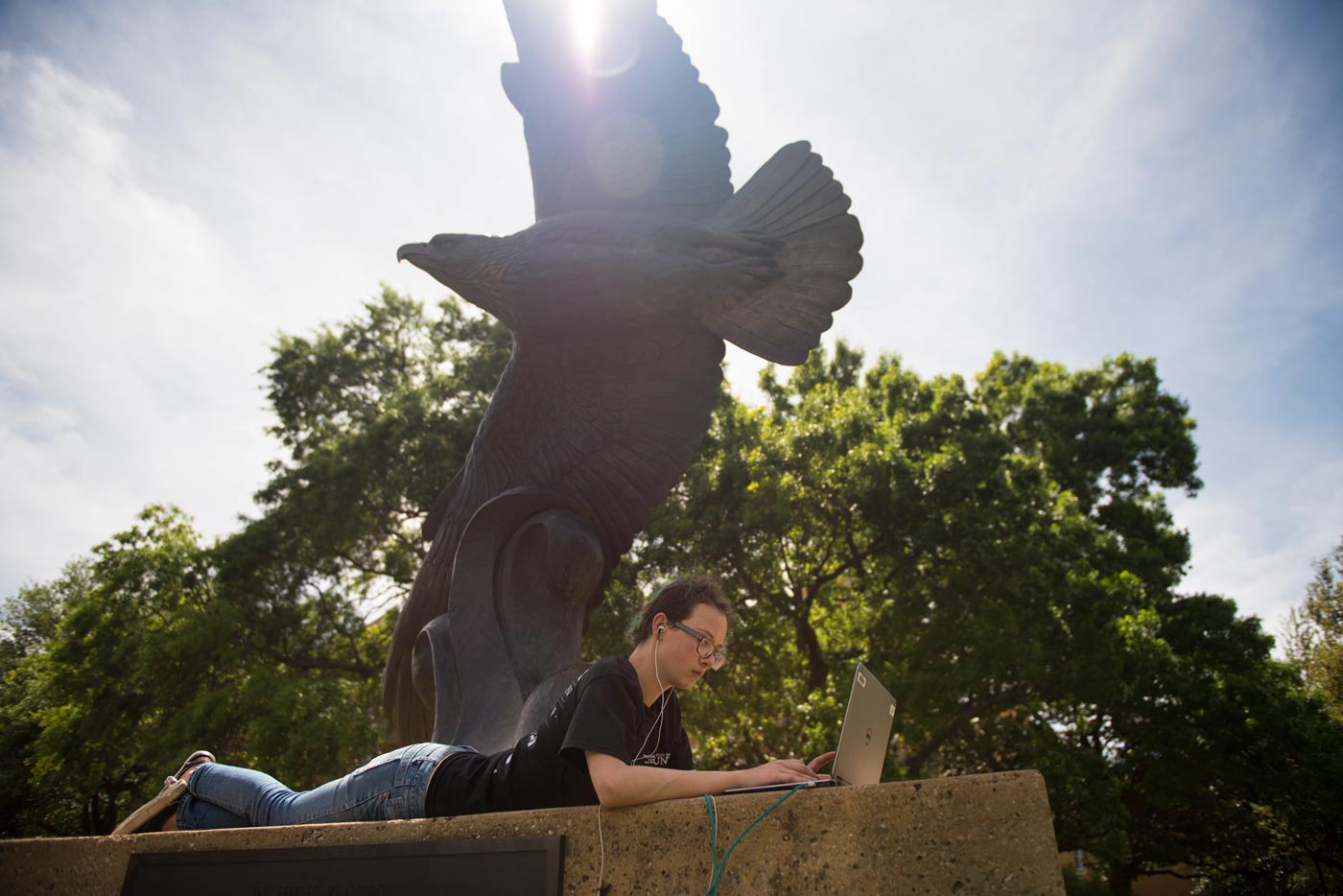 Student using a laptop in front of our eagle statue, with rays of sun light shining down.